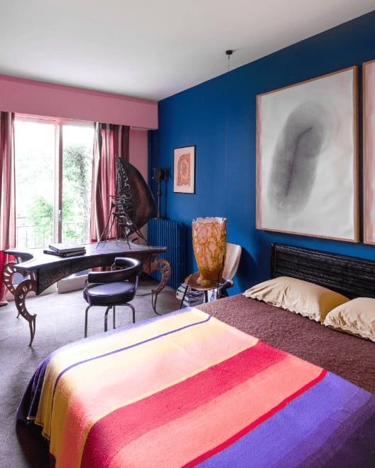 A black round chair sits at an eccentric desk in this eclectic bedroom showcasing carpet flooring and pink and royal blue wall mounted with large artworks.