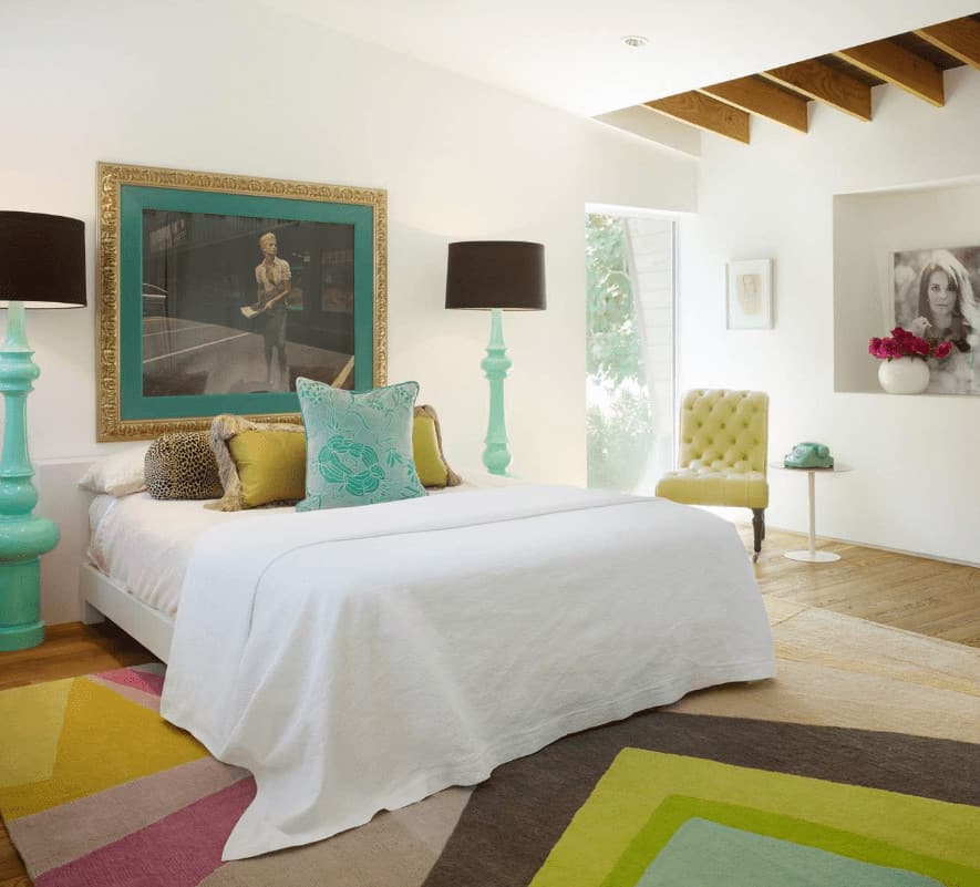 A tufted yellow green chair sits next to the white bed flanked by aqua floor lamps. This room has gorgeous framed photos and a striking printed rug that lays on the natural hardwood flooring.