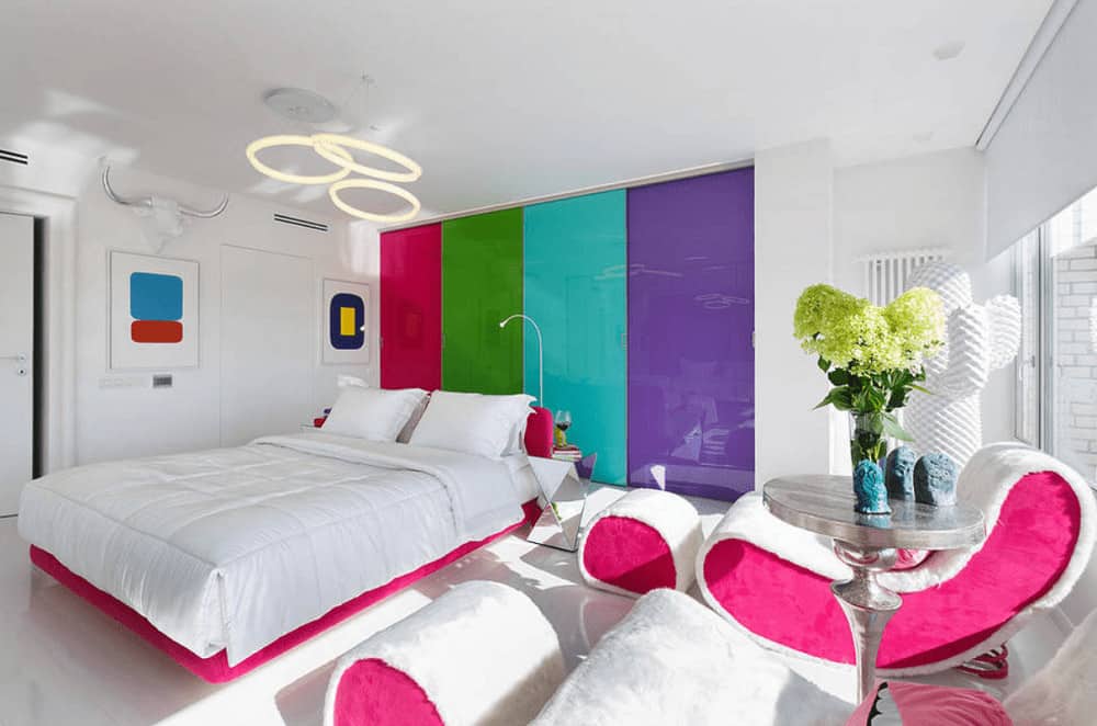 Stylish bedroom boasts white furniture with pink accents along with a built-in wardrobe fitted with multi-colored doors. It is illuminated by round pendant lights that hung over the platform bed.
