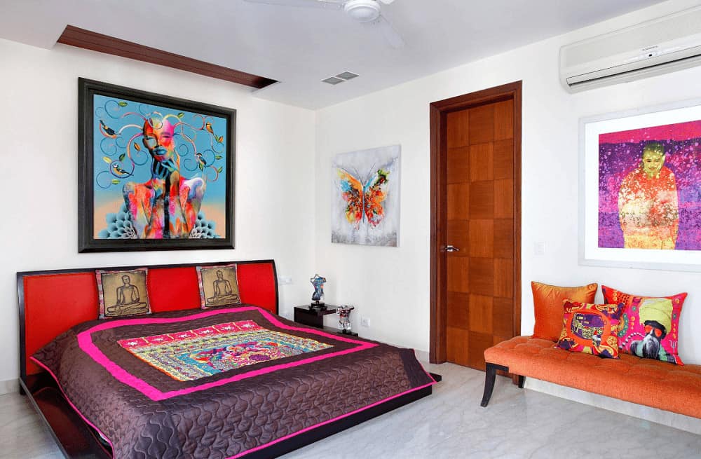 Eclectic bedroom offers an orange tufted bench and red platform bed paired with a dark wood nightstand. It is accented with gorgeous artworks mounted on the white walls.