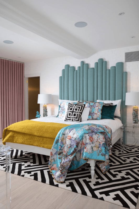 A blue floral throw blanket lays on the stylish bed in this primary bedroom with an eye-catching patterned rug and blush pink drapes covering the glazed windows.