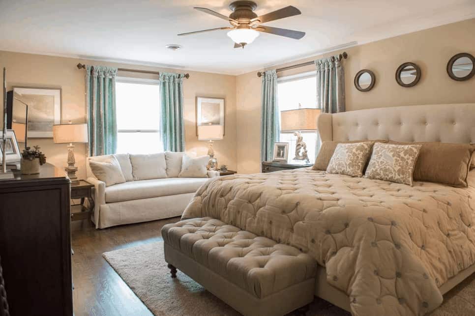 Beige primary bedroom with a skirted sofa and round wall arts that hung above the wingback bed with a tufted ottoman on its end. It has hardwood flooring and picture windows covered in charming blue draperies.