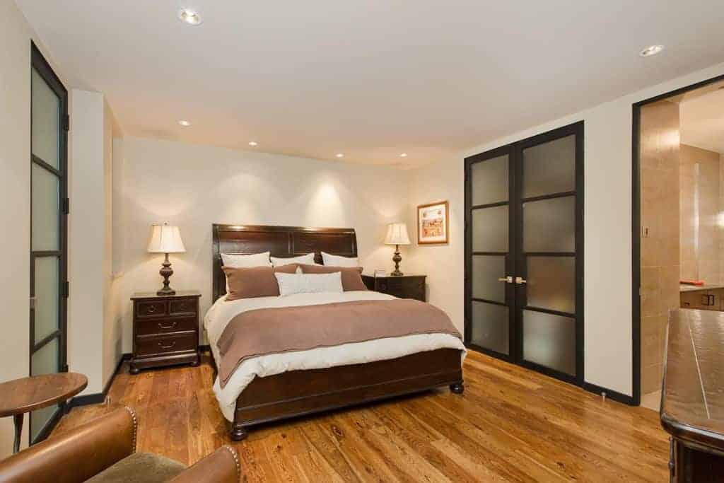 The cozy bedroom features a dark wood bed and matching nightstands over the rich hardwood flooring. It is lighted by traditional table lamps and recessed ceiling lights.