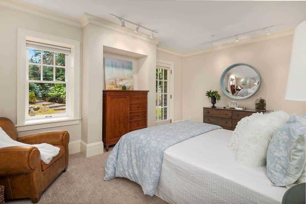 Airy bedroom decorated with a round concave mirror and lovely artwork that are illuminated by white track lights. It has a brown leather armchair and a comfy bed filled with fluffy and furry pillows.