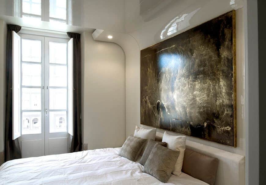 Natural light streams in through the glazed window in this primary bedroom with a cozy bed and interesting artwork mounted on the beige high gloss wall.
