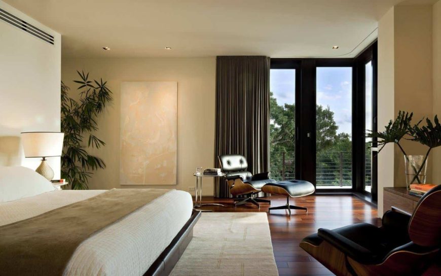 Fresh primary bedroom with black leather lounge chairs and a platform bed that sits on a beige area rug over wide plank flooring. It has full height glazing that overlooks the enchanting forest.