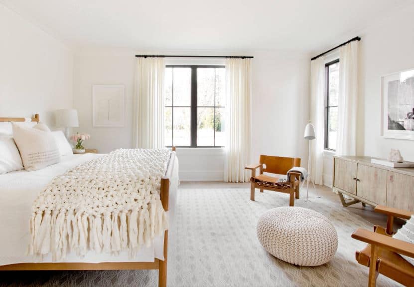 The wooden bed frame that is paired with a white cushion and sheet matches well with the pair of wooden armchairs at the sitting are by the foot of the bed. The sheets of the bed are as white as the walls and ceiling brightened by the windows.