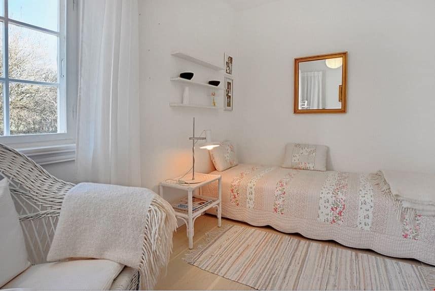 The simple bed is placed in a corner of white walls with side bearing floating white shelves and the other with a wall-mounted mirror. This is complemented by the light pink floral sheets of the bed that match the area rug on the light hardwood flooring.