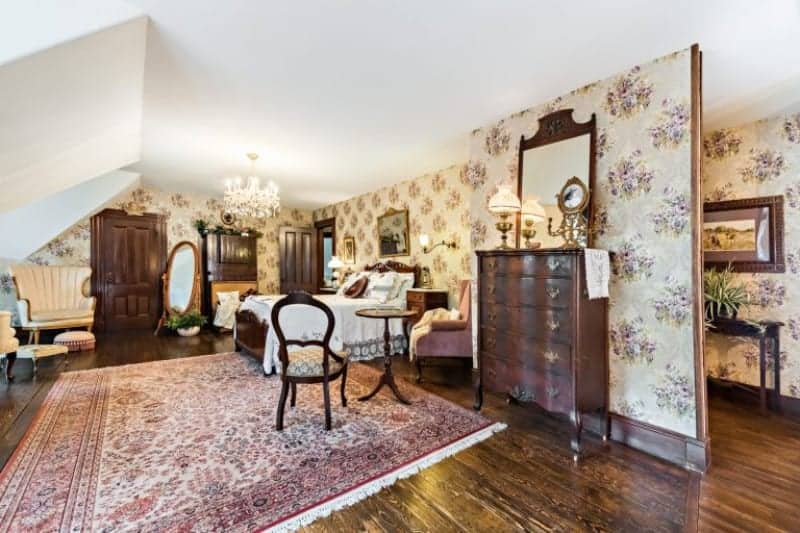 The lovely hardwood flooring is accented with a pink patterned area rug that extends from the sitting area to the wooden sleigh bed with white sheets and a wooden headboard that stands out against the beige floral wallpaper and white ceiling that has a brilliant chandelier.