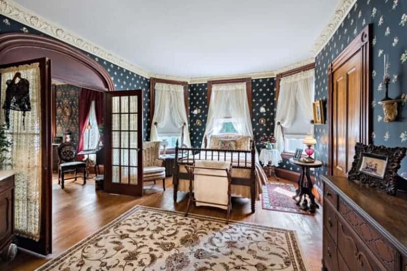 This gorgeous bedroom has a four-poster bed surrounded by curtained windows that brighten up the dark patterned wallpaper that dominates the walls. This also complements the wooden elements of the cabinets and hardwood flooring with floral area rug.