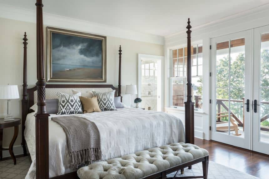 The dark brown pencil poster bed is the highlight of this Victorian-style primary bedroom with simple light beige walls and white ceiling. The light beige wall behind the cushioned headboard is accented with a beautiful painting of a cloudy beach.