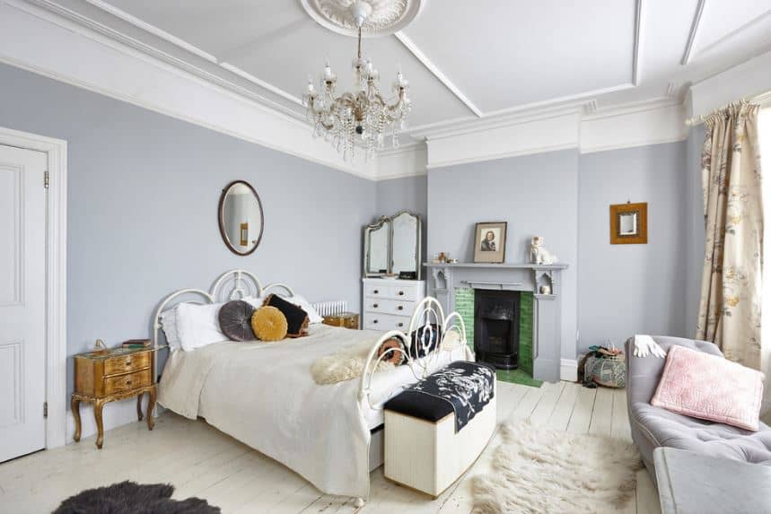 The light gray walls that go well with the white ceiling is accented with a fireplace by the side of the white wrought iron bed with matching white sheets. This is a nice complement to the white shiplap flooring illuminated by the brilliant windows by the foot of the bed.