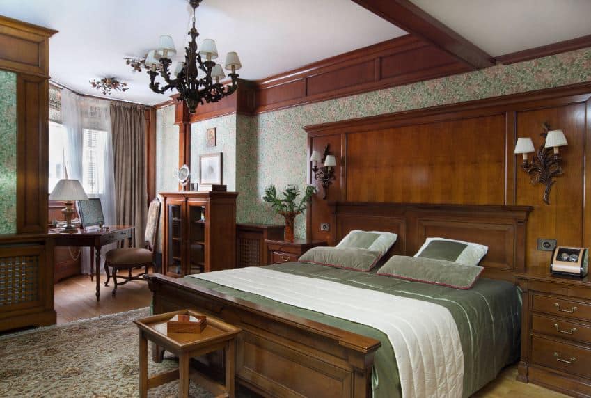 The intricate dark iron chandelier that stands out against the white ceiling is a match for the pair of wall-mounted lamps flanking the wooden headboard of the mission-style bed that has a pair of wooden bedside drawers blending with the wall panel.
