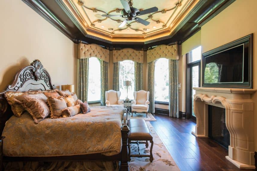 The beige mantle of the fireplace supports the TV across from the dark wooden sleigh bed with an elegant wooden headboard filled with carvings. This headboard contrasts the beige wall but matches with the ceiling and hardwood flooring.