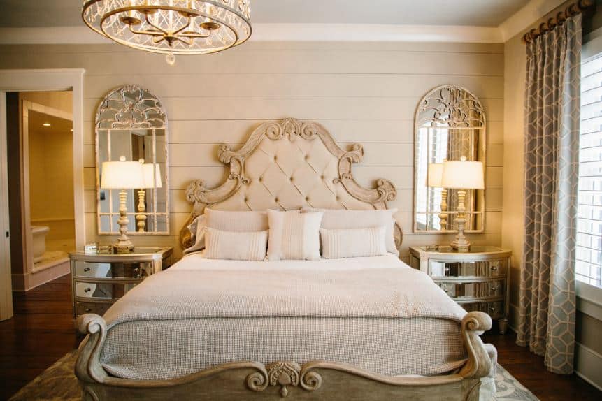 The two bedside drawers flanking the sleigh bed has a mirrored facade that matches with the wall-mounted mirrors above them. These mirrors stand out against the beige shiplap wall augmented by the warm yellow lights of the table lamps.