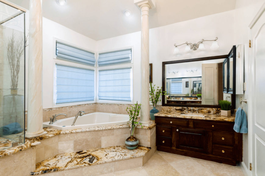 Mediterranean primary bathroom with tiled flooring and glazed windows covered in light blue roller blinds. It includes a dark wood vanity and corner tub lined with marble columns.