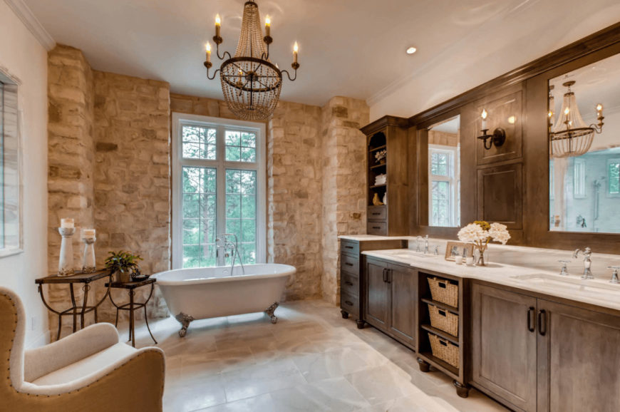 A stone brick wall adds texture in this primary bathroom with a cozy seat and clawfoot tub lighted by an oversized chandelier. It includes natural wood vanity with white marble countertop and dual sink.