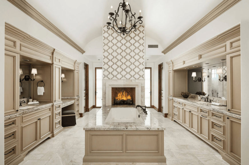 An ornate candle chandelier hangs over the drop-in tub attached to the patterned pillar that's fitted with a fireplace. It is flanked by built-in vanities with wrought iron hardware.