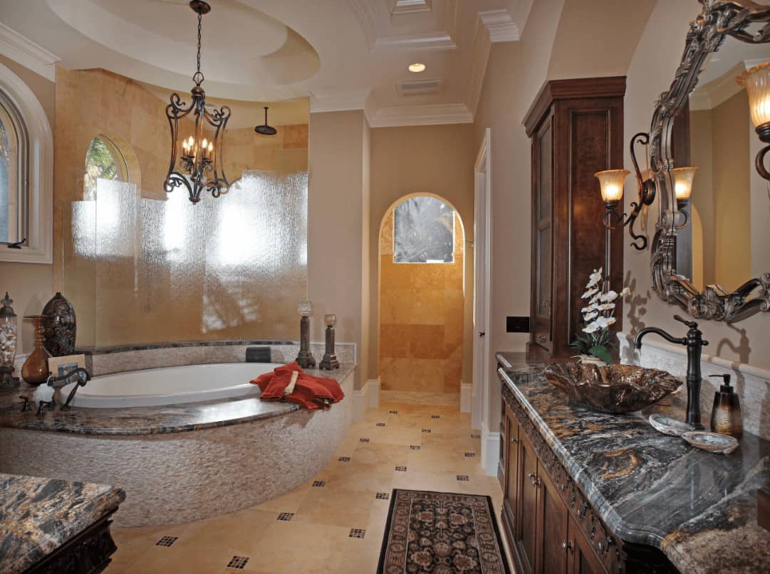A gorgeous carved wood mirror hangs above the wooden vanity that's topped with vessel sink and wrought iron faucet. There's a drop-in tub across lighted by an ornate chandelier that hung from the oval tray ceiling.