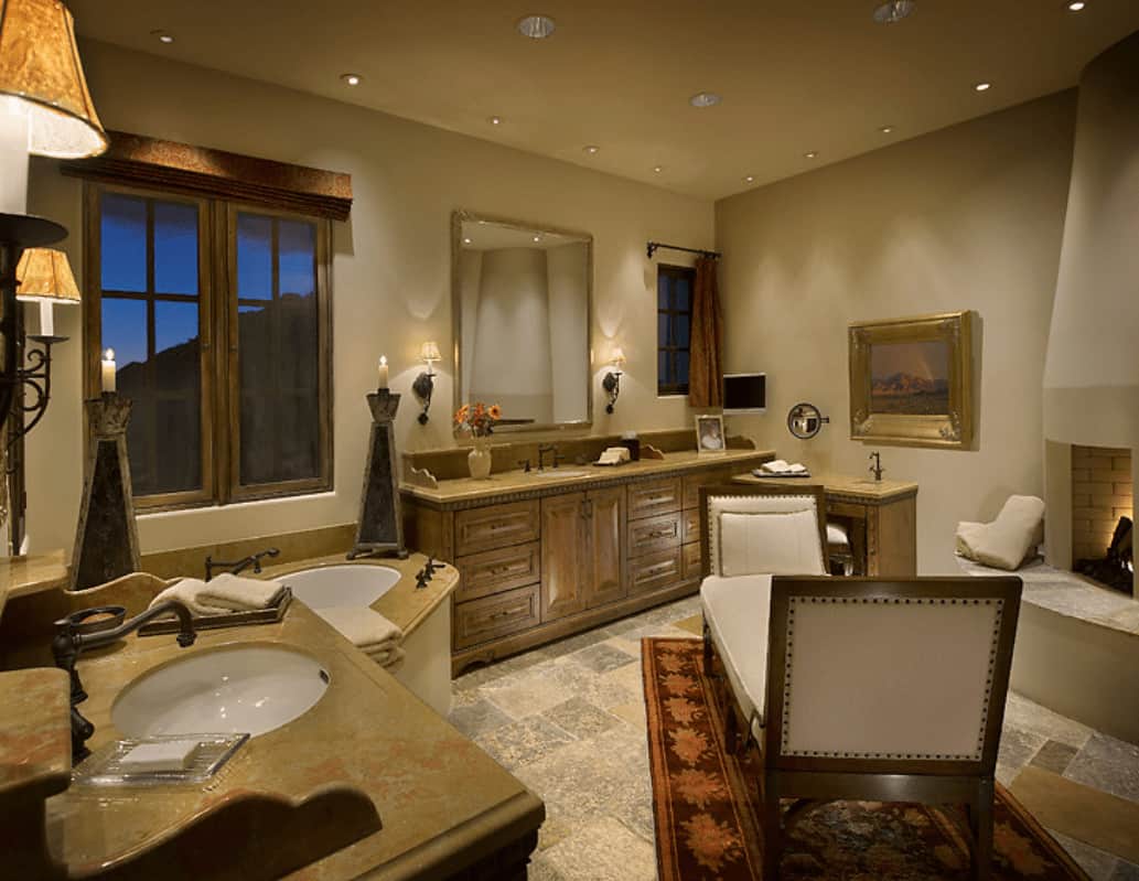 Natural wood vanities, drop-in tub and a kiva fireplace surround a beige cushioned bench that sits on a red bordered rug. This room is illuminated by wall sconces and recessed ceiling lights.