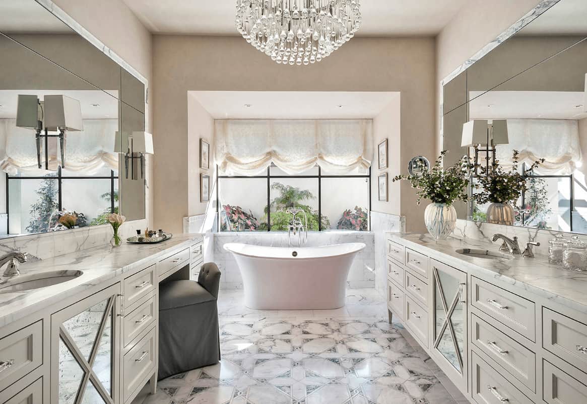 A fancy crystal chandelier adds class in this luxury primary bathroom with facing sink vanities and a freestanding tub by the framed window dressed in a charming roman shade.
