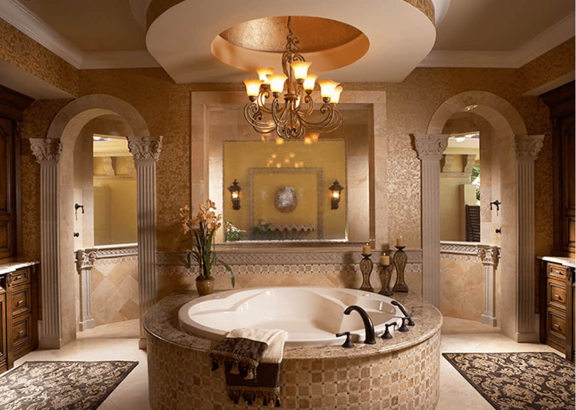 Elegant primary bathroom with a round bathtub illuminated by an ornate chandelier that hung from the tray ceiling. It includes classy rugs and arched doors leading to the walk-in shower.