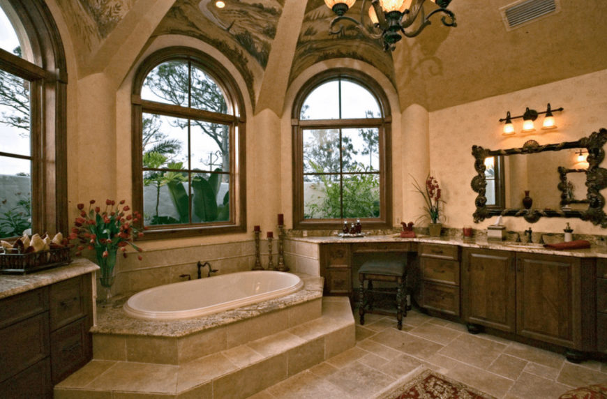 A green cushioned stool sits at a wooden sink vanity paired with a carved wood mirror. This room has limestone flooring and arched windows allowing natural light in.
