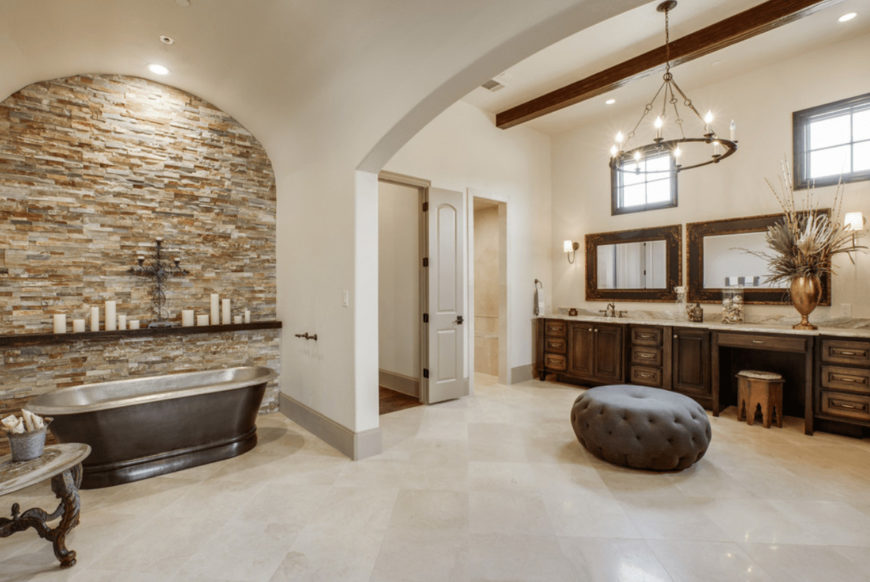Spacious primary bathroom offers a freestanding bathtub and wooden sink vanity accompanied by a gray tufted ottoman that's lighted by a round candle chandelier.