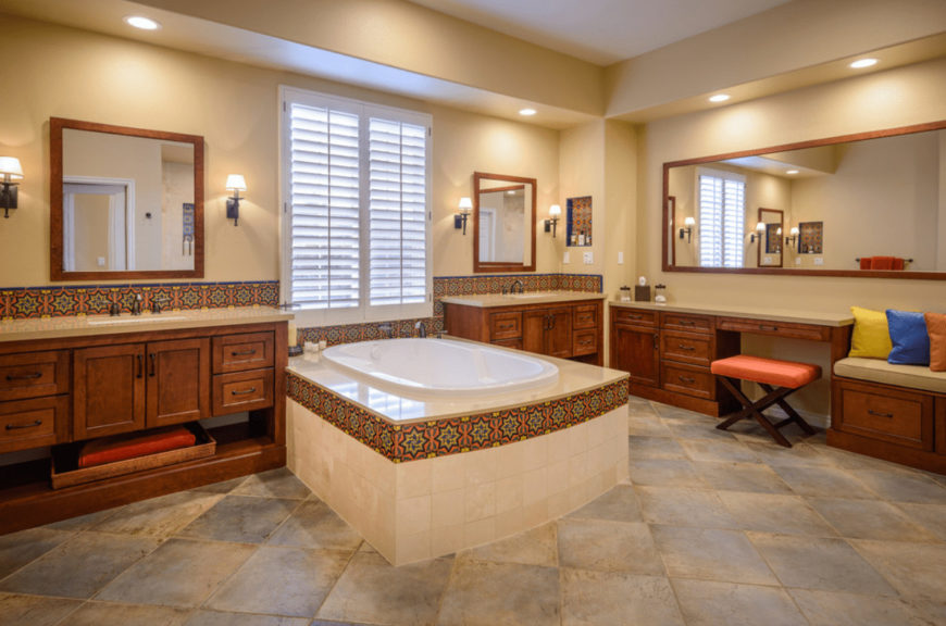 The Mediterranean primary bathroom features a deep soaking tub and wooden vanities lined with eye-catching decorative tiles. There's a built-in seat on the side topped with beige cushion and multi-colored pillows.