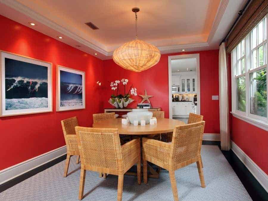 The white tray ceiling has a warm yellow glow to its borders that is augmented by the pendant light with a rustic hood that matches the dining chairs that have woven wicker designs. All of these are contrasted by the bright red walls accented with framed photos of surfers riding a large wave.