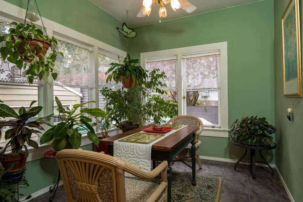 The various potted plants that dominated this Tropical-style dining room is complemented by the green walls brightened by the white frames of the windows that bring in natural lighting for the simple wooden dining table and its two bamboo armchairs that stand out against the gray flooring.