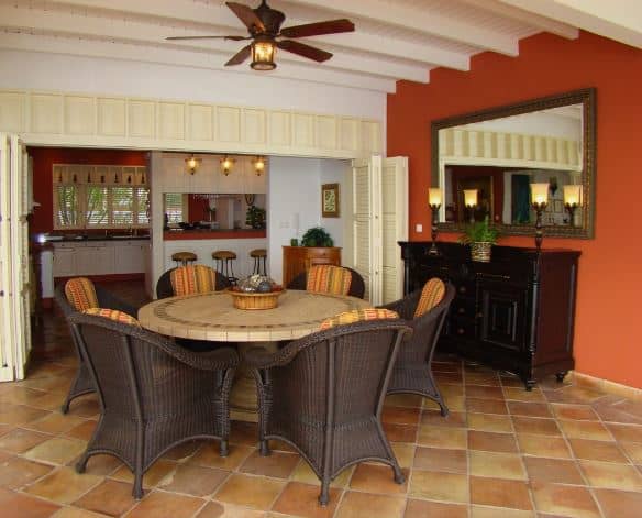 The earthy terracotta flooring tiles of this Tropical-style dining room complements the dark brown elements of the cabinet and the woven wicker dining chairs. It also complements the red wall that contrasts the white wooden ceiling with a dark brown ceiling fan in the middle.