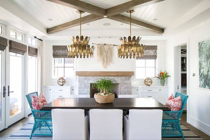 The bright white ceiling is complemented by two exposed wooden beams in a cross design. There is also a pair of decorative pendant lights with leaf designs hanging over the dark wooden dining table that has two kinds of dining chairs. There are two rustic green bamboo chairs and armchairs with white slipcovers.