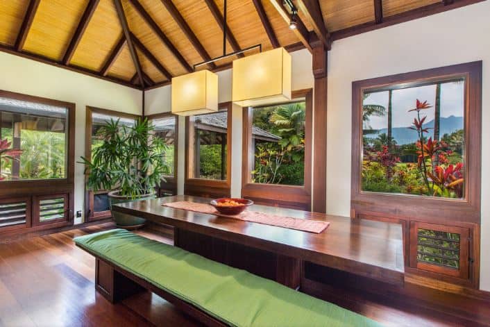 The highlight of this Tropical-style dining room is the lush green scenery of the landscaping outside that is featured by the surrounding windows. These windows have wooden frames that match the hardwood flooring, dining table and its wooden benches with green cushions on them.