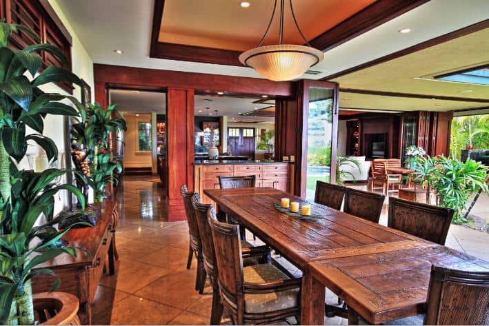The terracotta flooring tiles are a good match for the wooden dining table and its wooden chairs with a burnt design on its hue. This palette is mirrored by the tray ceiling with dark wooden borders and a dome pendant light with a shell design.