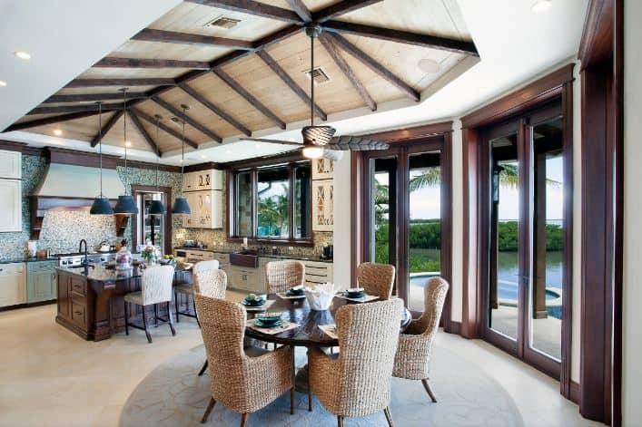 This informal dining room is in the same large room as the kitchen. They are both under a wooden cathedral ceiling with exposed beams supporting a ceiling fan with lighting that has a leaf design. This hangs over the round wooden dining table that stands out against its woven wicker chairs and the beige carpeted flooring.