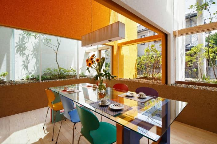 This informal dining room has modern chairs of different colors that complement the modern glass-top dining table. This is surrounded by tall glass windows with plant boxes that have plants just outside the glass for that unique tone to pair the orange and brown textured walls.
