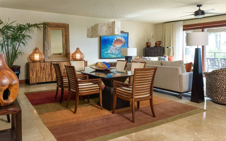 This dining area is right beside the living room area that has light gray sofas and a brilliant blue painting of a seashell. This painting adds to the adornment of the beige walls like the wooden decors, mirror and table lamps with a rustic seaside vibe. These serve as background for the modern dining table with a glass top and its wooden slat chairs.
