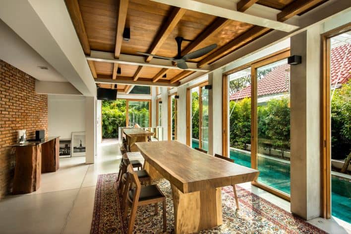 The large white tiles of the flooring is topped with a colorful patterned area rug that gives contrast to the light wooden dining table paired with wooden chairs that have black leather cushioned seats. This setup has a nice view of the blue pool just outside the glass sliding doors.