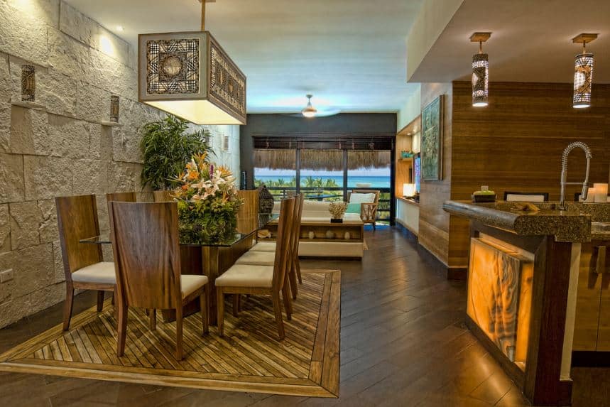 This informal dining room is beside the kitchen bar has a lit frosted glass panel on its front. The dining area is marked by a change in the dark hardwood flooring beneath the wooden dining table and the wooden chairs with beige cushioned seats. This matches with the decorative rectangular pendant light over the glass top of the table.