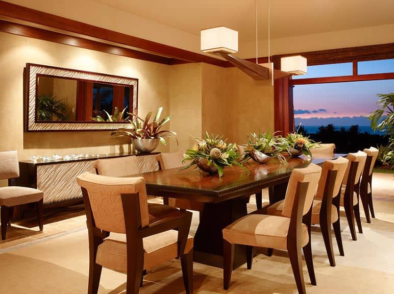 There is a decorative pendant light hanging from the beige ceiling that has two hood lights on each end. The beige ceiling matches the walls that are adorned with a wall-mounted mirror with frames matching the facade of the dining room cabinet below it. They are complemented by the large dining set of wooden table and wooden chairs with beige cushions.