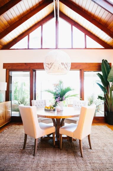 The brightness of the natural lights coming in from the large glass doors and transom windows makes the white hood of the pendant glow as well as the white leather cushioned dining chairs. This brightness is balanced with the dark wooden elements of the cathedral ceiling, round table and frames of the doors and windows.