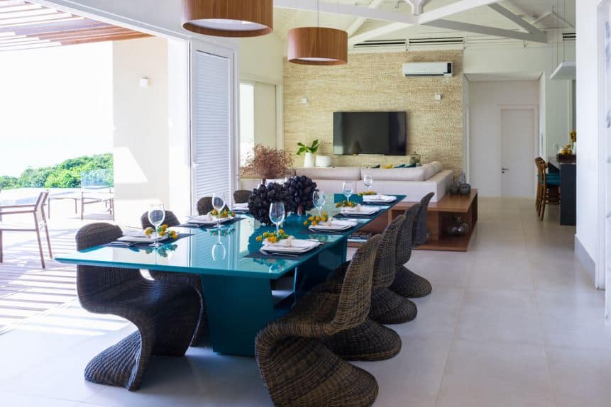 The modern blue rectangular dining table is surrounded by rustic woven wicker chairs that stand out against the white tiles of the floor that is illuminated by the natural lights coming from the large entryway from the yard.