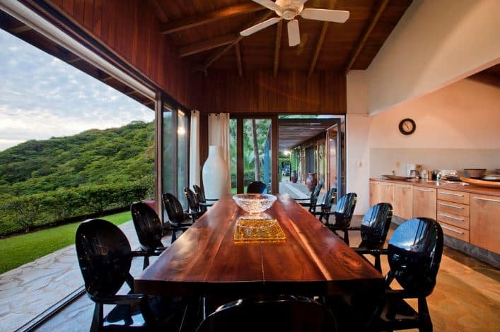 A whole wall of this Tropical-style dining room is open to the lush green landscaping outside through large glass sliding doors that bring in abundant natural lighting to the dark wooden elements of the ceiling with exposed wooden beams and dining table with modern black chairs.