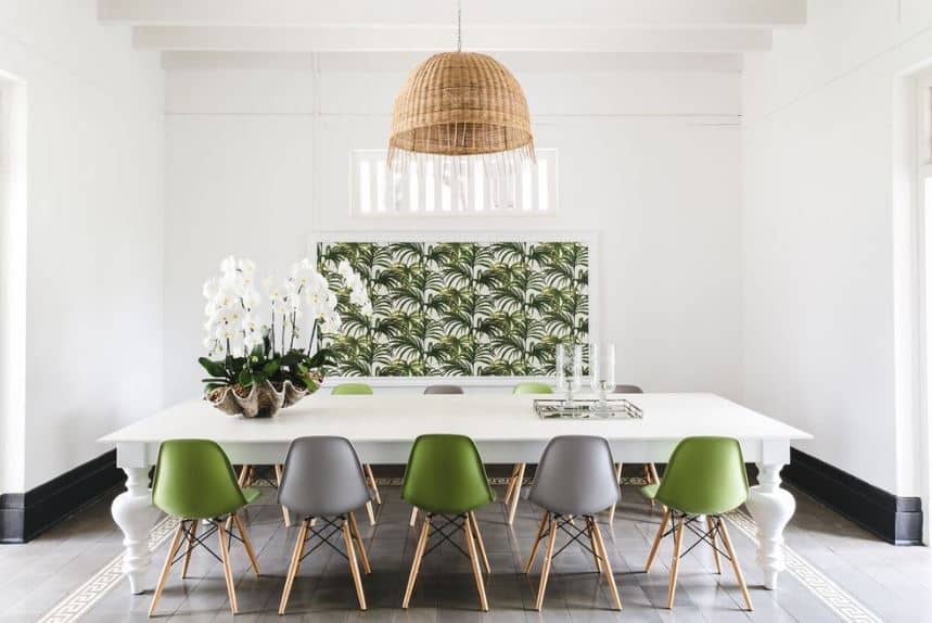 The brown rustic basket hood of the dome pendant light stands out against the white ceiling with exposed wooden beams and white walls as well as the white dining table. These are contrasted by the green elements of the modern dining chairs and the framed artwork of leaves paired with a potted plant in a large shell.