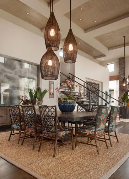Three pendant lights and their rustic woven wicker hoods pairs well with the brown woven area rug over the dark hardwood flooring. This matches the dark wooden hue of the rectangular dining table and its bamboo chairs with colorful floral cushions.