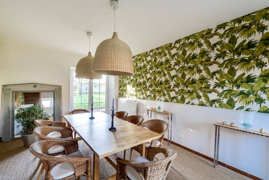 The long rectangular wooden dining table has a light hue to it complemented by the woven wicker armchairs with white seat cushions. These chairs blend with the brown carpeted flooring and the two large rustic basket hood of the pendant lights.