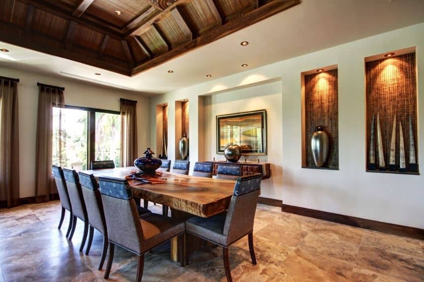 The earthy wooden ceiling is paired with contrasting white walls that have small alcoves on it for decor display that has recessed lights. The large rectangular dining table matches the marble flooring and the brown cushioned dining chairs surrounding it.