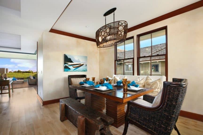 The rustic wooden bench that is made of a single thick beam matches well with the rustic dark woven wicker chairs at each end of the wooden dining table that is topped with a decorative lighting with intricate patterns on it that contrasts the white ceiling.