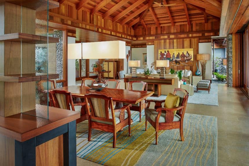 The large wooden cathedral ceiling has wooden exposed beams that support the rectangular pendant light hanging over the redwood dining table and its wooden armchairs with light gray cushions that work well with the blue patterned area rug over the gray flooring.